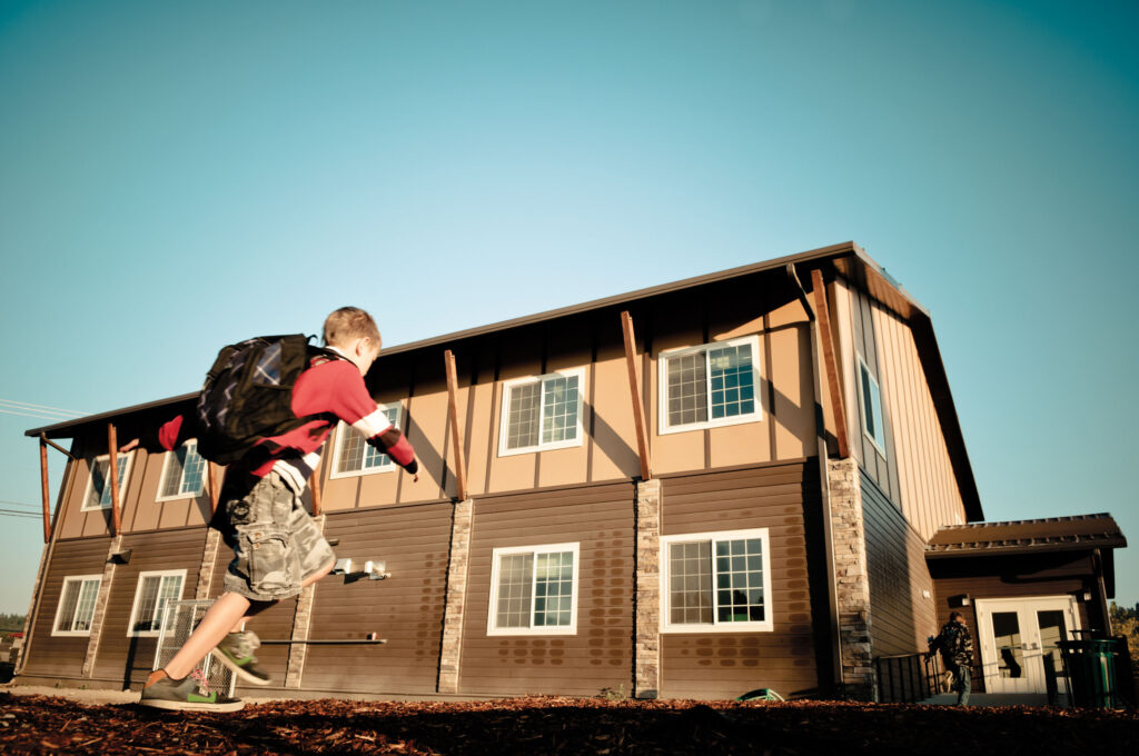 A boy in a red shirt & backpack runs towards a brown building