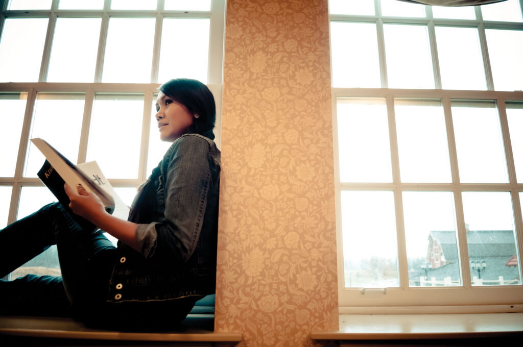 A woman sitting on a windowsill reading a book with a barn in the background