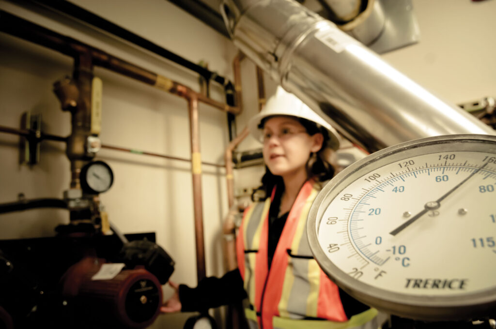 A worker standing with a hardhat and high visibility vest with pipes in the background
