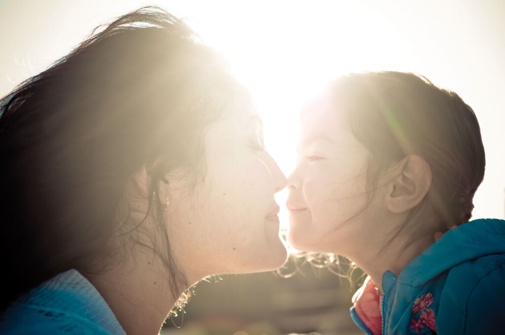A woman and child face each other with noses touching and the sun shines in the background.